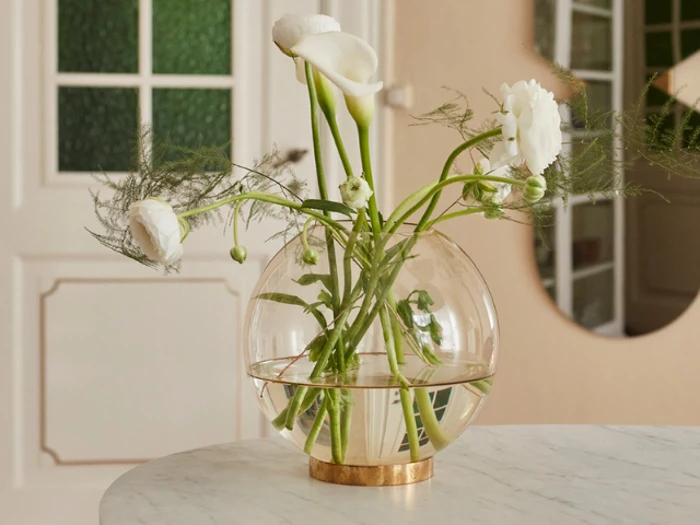 En rund glasvase med hvide blomster og grønne stilke står på et marmorbord i et blødt oplyst, moderne rum.