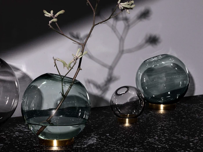 Tre runde glasvaser med guldbund står på en mørk plettet overflade; i den ene vase står en tynd gren, og bløde skygger falder på den lysegrå væg bag dem.