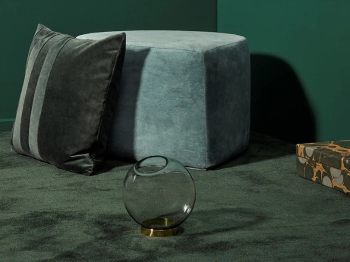 En rund grå ottoman, en stribet grå pude, en rund glasvase med guldfod og en mønstret æske på et mørkegrønt tæppe mod grønne vægge.