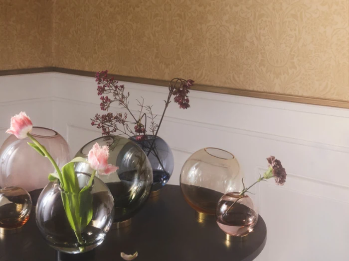 Flere runde glasvaser, nogle med lyserøde blomster og andre tomme, står på et mørkt bord mod en beige og hvid væg.