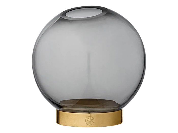 En rund, røggrå glasvase står på en flad, cirkulær base af guldmetal. Vasen har en bred krop med en lille åbning i toppen.