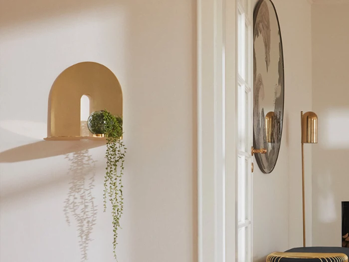 En væghylde i guld med en glaskugle og en slynget plante ved siden af en hvid dør og en rund vægkunst i et lyst, moderne rum.