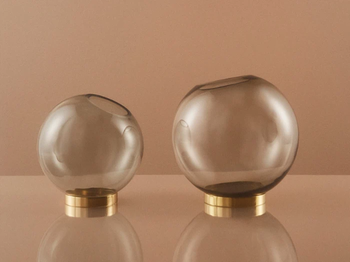 To kugleformede glasvaser med asymmetriske åbninger står på guldbaser mod en beige baggrund og reflekterer lyset på den blanke overflade.