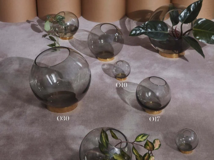 Glasboblevaser i forskellige størrelser, nogle med grønne planter, udstillet på en lyslilla overflade med solbrune cylindriske genstande i baggrunden.