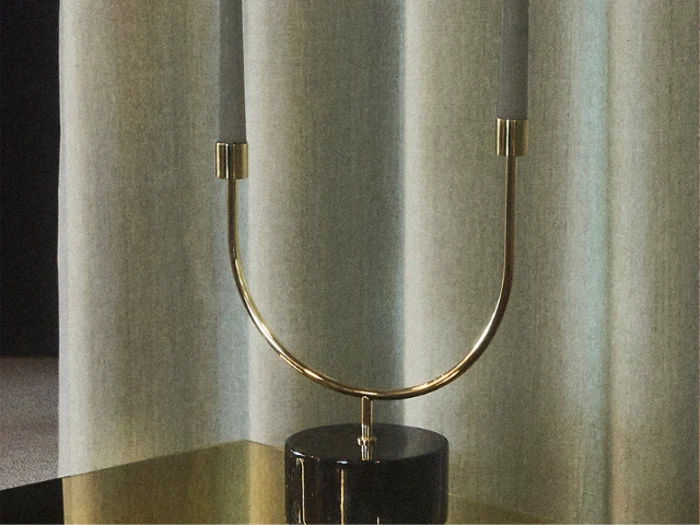 En moderne, U-formet guldlysestage med to hvide lys står på en reflekterende overflade foran lyse gardiner.