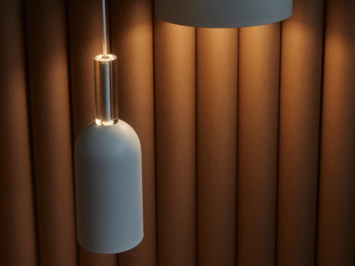 En moderne pendel hænger foran en brun, lodret polstret væg, der kaster et varmt skær og skaber en hyggelig, stilfuld stemning.