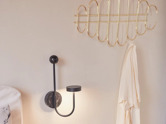 En moderne væglampe med en rund, sort base og en buet arm er monteret på en lys væg ved siden af et guldfarvet vægstativ med et cremefarvet stykke tøj.