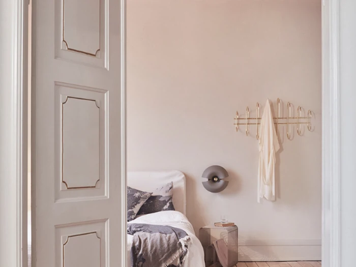 Et minimalt soveværelse med en hvid seng, et tæppe, et lille sidebord, en væglampe og guldkroge, der holder et hængende hvidt stykke tøj.