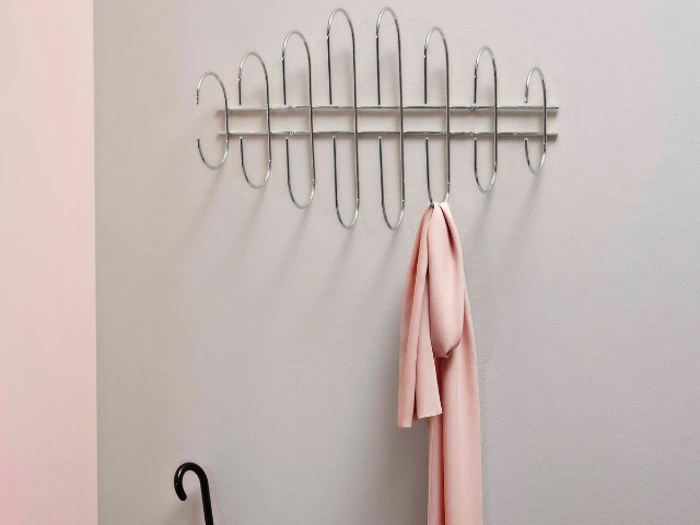 Et moderne vægstativ i metal med et abstrakt ovalt design holder et lyserødt tørklæde; en sort paraply læner sig op ad den lyse væg nedenunder.