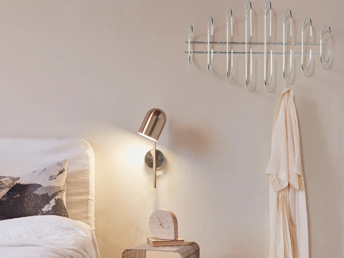 Et moderne soveværelse med en væghængt metallisk knagerække, en beige morgenkåbe, en guldlampe og et lille natbord med et ur og en bog.