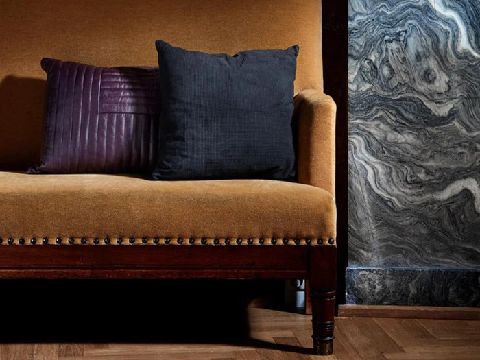 En sennepsgul polstret sofa med træben står foran en marmoreret væg. To puder, en mørklilla og en sort, hviler på sofaen.