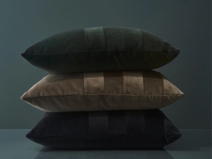 Tre fløjlspuder i mørkegrøn, brun og sort er stablet oven på hinanden mod en mørk, minimalistisk baggrund.