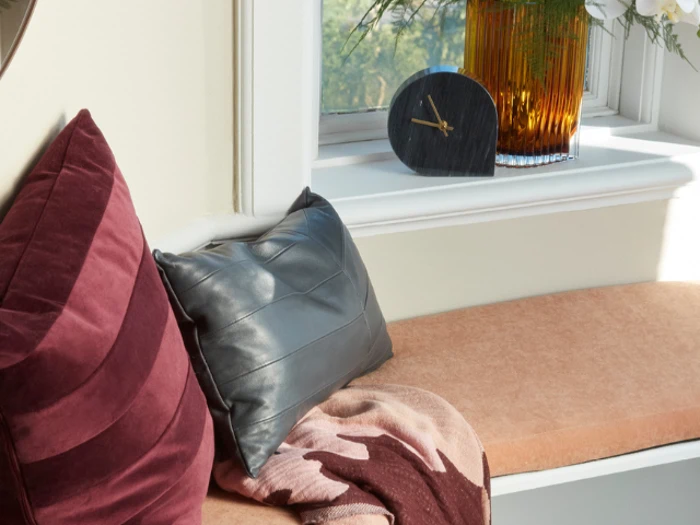 Et hyggeligt vinduessæde med en lilla pude, en grå pude, et mønstret tæppe, et lille ur og en vase med blomster i vindueskarmen.