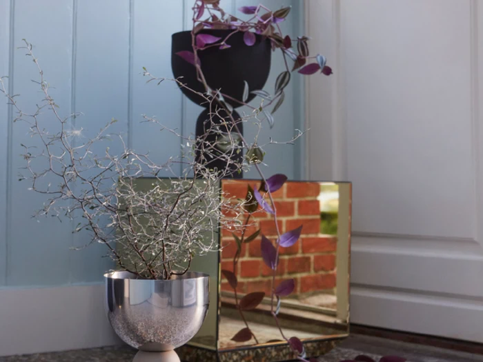 En sølvpotte med en bladplante står foran et spejl, der reflekterer en murstensvæg; en anden plante i en sort potte er placeret bagved, med lilla slyngplanter.