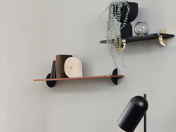 To minimalistiske væghylder med bøger, pyntegenstande, en potteplante og en rund lampe, monteret på en lysegrå væg over en sort skrivebordslampe.