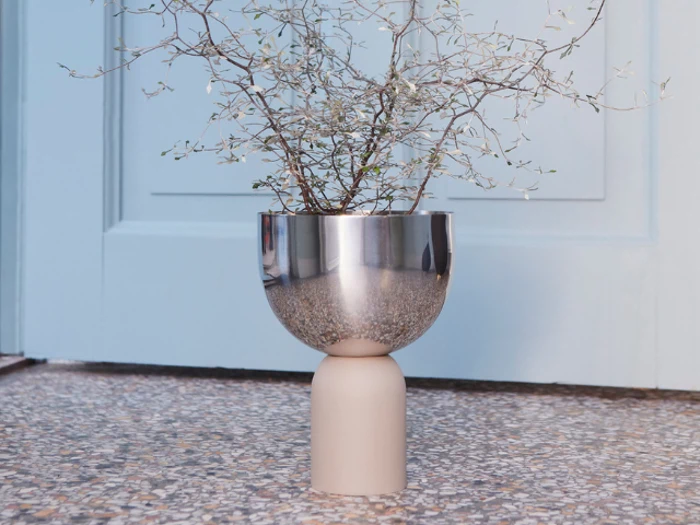 En minimalistisk plante med tynde grene står i en moderne, reflekterende vase i metal og beige på et spættet stengulv foran en lyseblå dør.