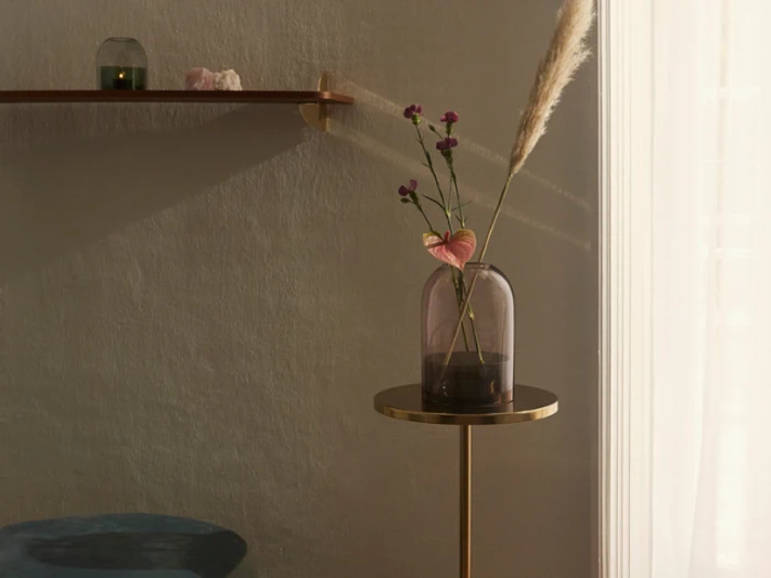 En lilla glasvase med pink og lilla blomster står på et lille rundt bord ved siden af et svagt oplyst vindue; på væggen ovenover står en hylde med dekorationer.