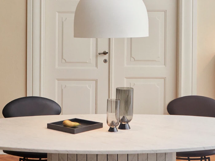 Rundt hvidt bord med to grå glas, en sort bakke med en enkelt gul frugt, to sorte stole og en stor hvid pendellampe over. Hvide dobbeltdøre ses i baggrunden.