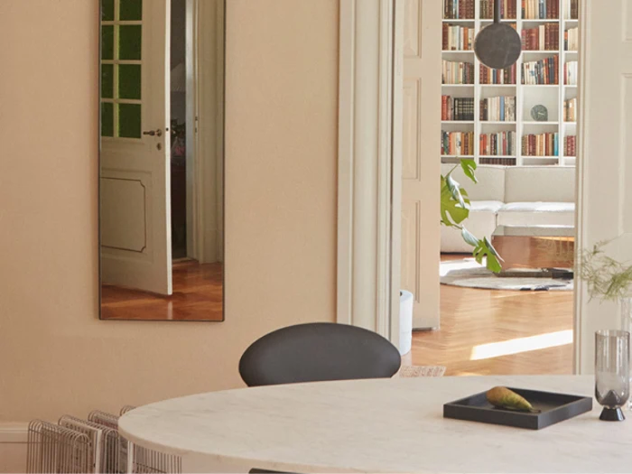 Et moderne, solbeskinnet rum med et rundt bord og en sort stol i forgrunden, et højt spejl på væggen og et hyggeligt opholdsområde med bogreoler og en hvid sofa i baggrunden.