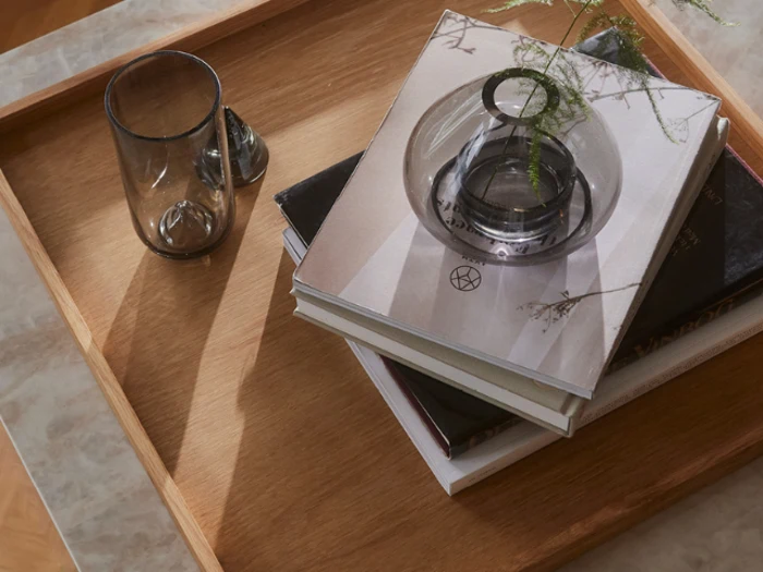 På en træbakke står et glas, en lille vase med en plantestilk og en stak bøger på en marmorplade.