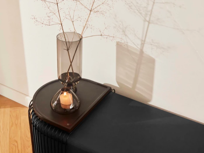 En lille glasvase med tynde grene og et tændt stearinlys står på en mørk bakke oven på et moderne sort skab mod en hvid væg.