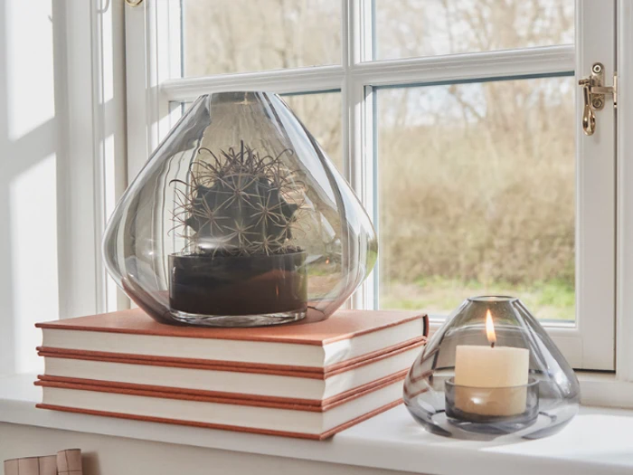En glasvase med en kaktus indeni står på stablede bøger ved et vindue; ved siden af står et tændt stearinlys i en matchende glasholder.