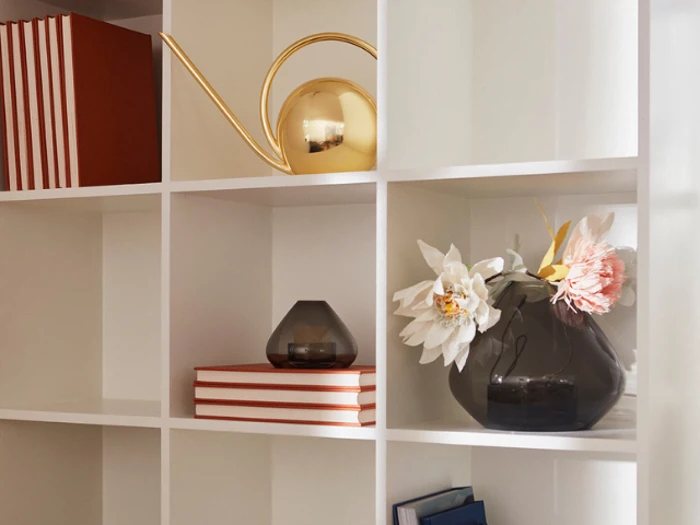 Hvid reol med stablede bøger, en guldvandkande, en mørk glasvase med blomster og en lille mørk glasbeholder, alt sammen pænt anbragt i hver sin kasse.