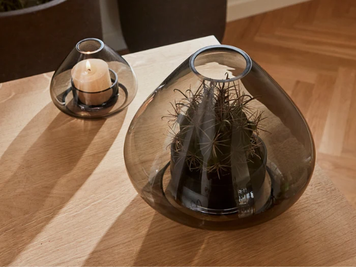 To glasvaser står på et træbord; den ene indeholder et stearinlys, den anden en lille kaktus. Begge vaser har et røgfarvet, gennemskinneligt udseende.
