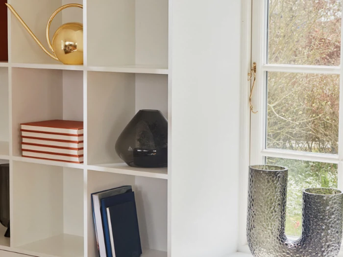 Hvid reol med bøger, en sort vase og en guldvandkande. Ved siden af er der et vindue med to teksturerede glasvaser i vindueskarmen og træer, der kan ses udenfor.