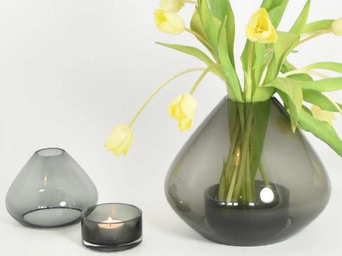 Der vises tre røgfarvede glasvaser; en indeholder gule tulipaner, en er tom, og den mindste indeholder et tændt fyrfadslys, alt sammen mod en hvid baggrund.