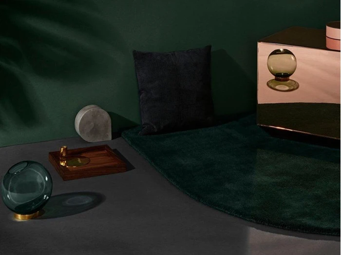 En moderne interiørscene med et mørkegrønt tæppe, en sort pude, en rund glasgenstand, en betonindretning, en træbakke og et spejlskab mod en mørkegrøn væg.