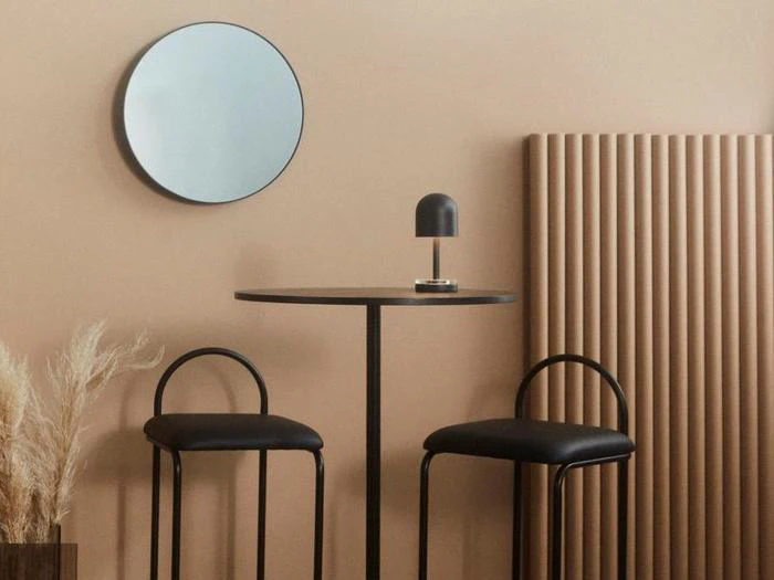 Minimalistisk indretning med et rundt vægspejl, et lille sort bord, to sorte stole, en lille lampe, en beige væg, tørret pampasgræs og et lodret, riflet panel.