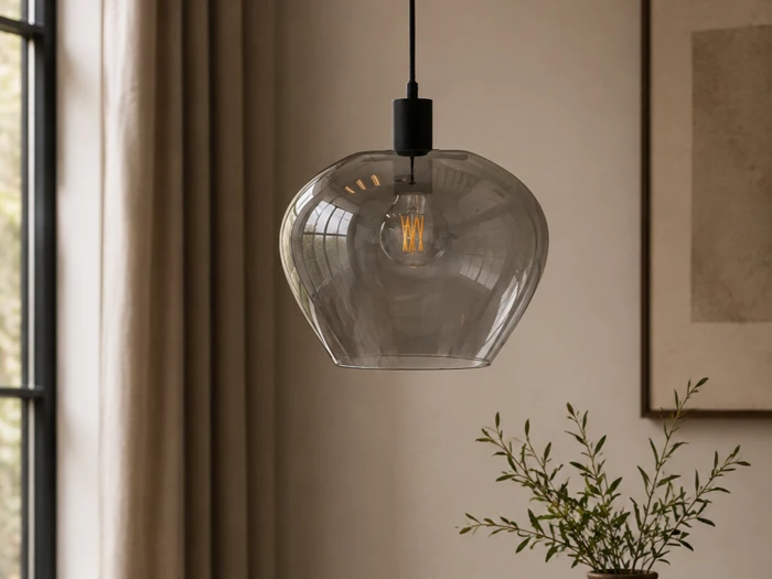 En pendel i klart glas med en pære med synlig glødetråd hænger i et blødt oplyst rum tæt på et vindue med beige gardiner og en plante i forgrunden.