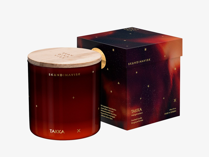 Et rødt glaslys med et trælåg mærket SKANDINAVISK TAKKA står ved siden af en matchende mørk, stjernemønstret æske.