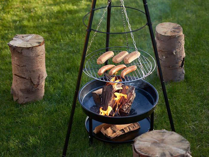 Pølser grilles over åben ild på en trebenet grill i en græsklædt gård med træstubbe som siddepladser omkring bålet.