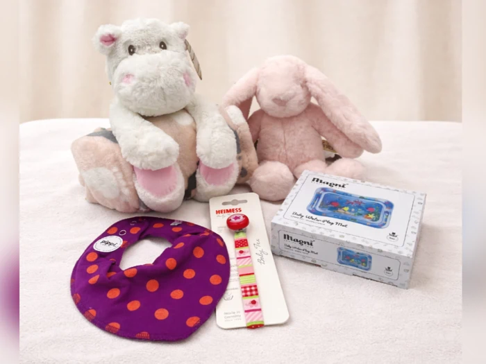 Plyslegetøj med flodhest og kanin, en lilla, prikket hagesmæk, en sutteklips og en vandlegemåtte i æske udstillet på en lys overflade.