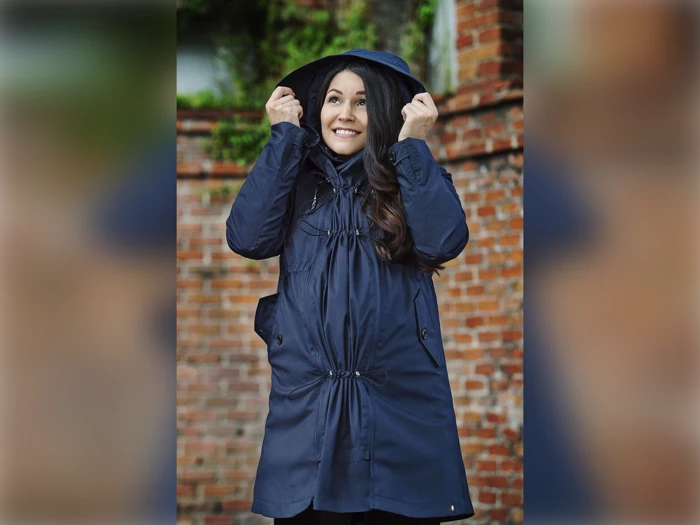 En smilende kvinde i en marineblå regnfrakke holder sin hætte op, mens hun står udenfor foran en murstensvæg med grønne planter.
