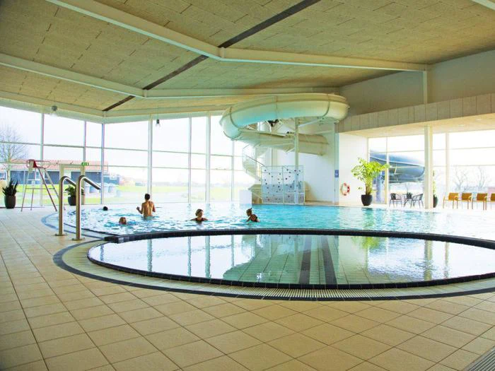 Indendørs swimmingpool med vandrutsjebane, flere personer i vandet, store vinduer, der lukker naturligt lys ind, og synlige siddepladser ved poolen.