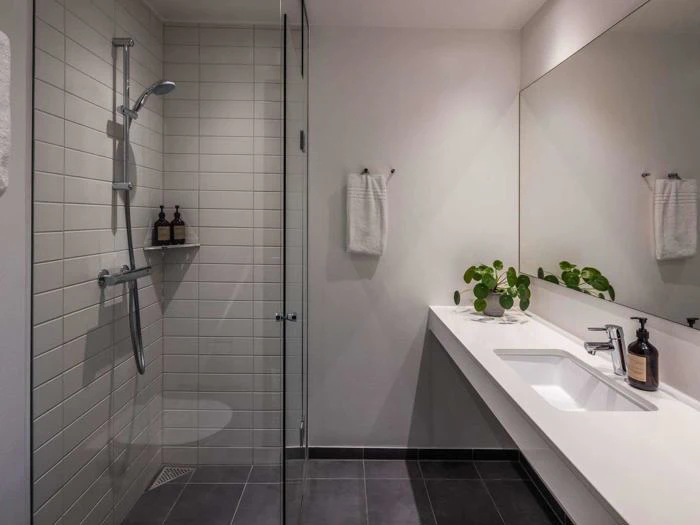 Moderne badeværelse med glasbruser, hvide flisevægge, gråt gulv, lang hvid bordplade med vask, stort spejl, potteplante og sæbedispensere.