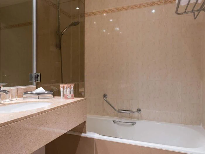 Et beigefarvet badeværelse med badekar, væghængt bruser, støtteben og et bord med toiletartikler, en håndvask, servietter og et glas. Håndklæderne er foldet på et stativ over badekarret.