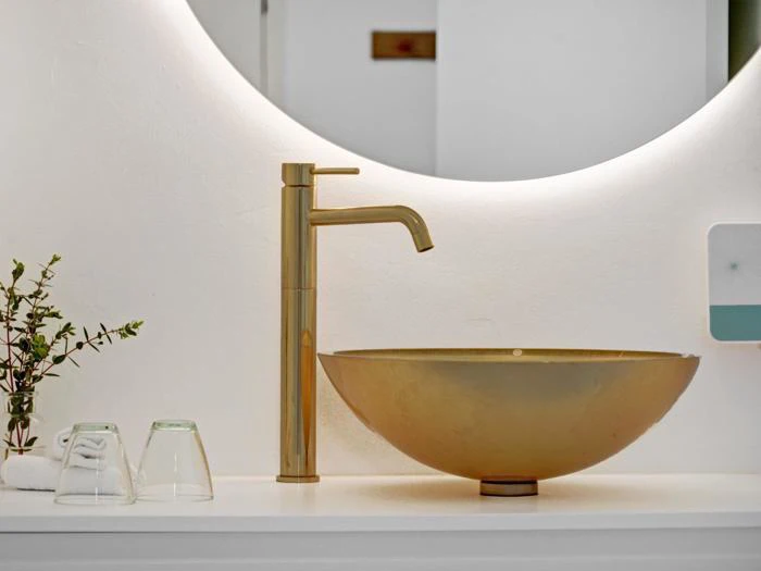 Moderne håndvask med en rund guldvask og matchende vandhane, to glaskopper, et sammenrullet håndklæde og en lille plante på en hvid baggrund med et rundt spejl.
