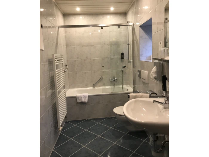 Moderne badeværelse med flisebelagte vægge og gulv, badekar med glasdøre, væghængt håndvask, toilet, håndklædetørrer og et lille vindue over badekarret.