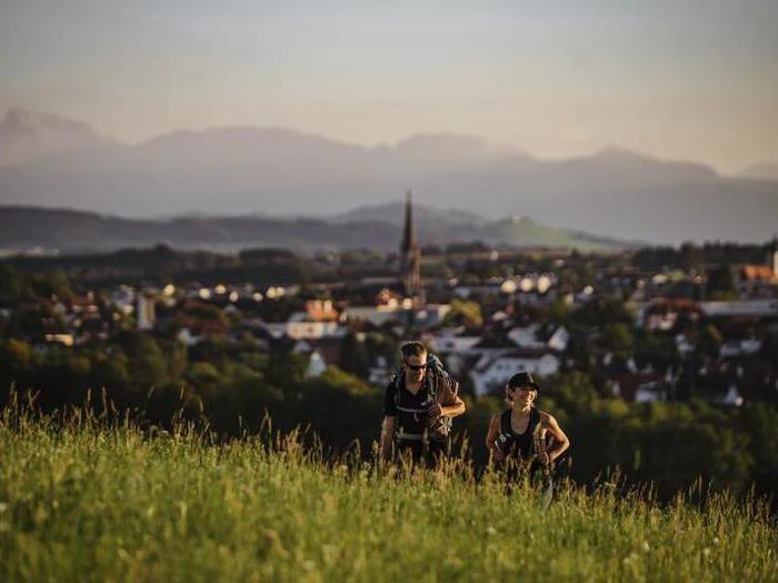 To mennesker vandrer op ad en græsklædt bakke med en by og et kirkespir i baggrunden, og bjergene ses i det fjerne under en klar himmel.