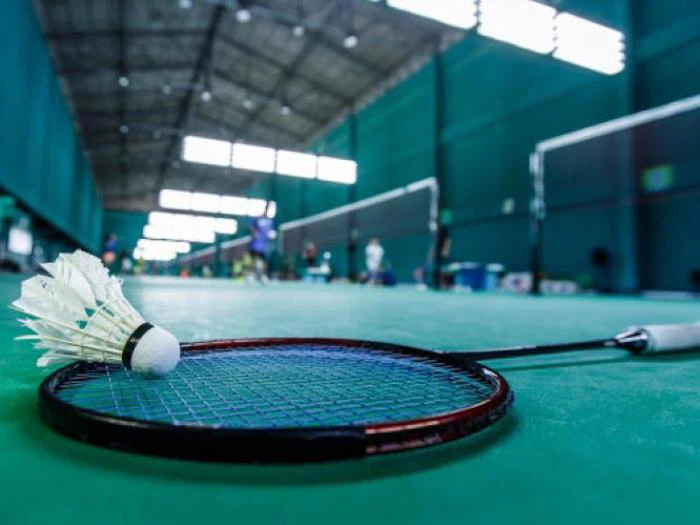 En badmintonketcher og en fjerbold hviler på en grøn indendørs bane med slørede spillere og net i baggrunden.