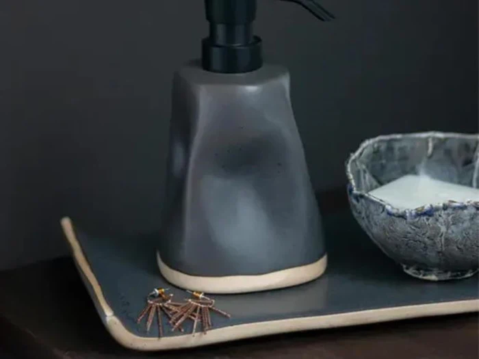 En matgrå sæbedispenser står på en bakke ved siden af en lille struktureret skål og et par øreringe.