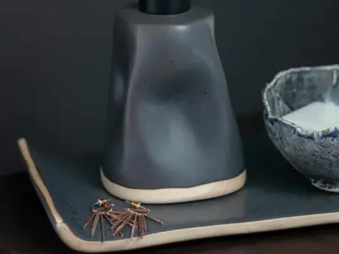 Et par edderkoppeformede øreringe ligger på en mørk bakke ved siden af en keramisk genstand og en struktureret skål.