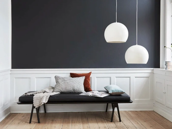 Et minimalistisk rum med en sort bænk, forskellige puder, et åbent magasin, to hvide pendellamper og vægge beklædt med mørkegrå og hvide paneler.