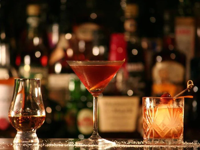 Tre cocktails - en whisky neat, en rød martini og en old fashioned med kirsebær - står på en bardisk med slørede flasker i baggrunden.