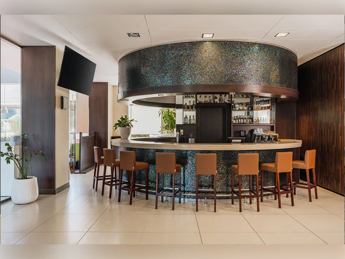 Moderne cirkulær hotelbar med brune barstole, mosaikfliser foran og hylder med flasker bag disken. Store potteplanter står på hver side af baren i et lyst, stilfuldt rum.