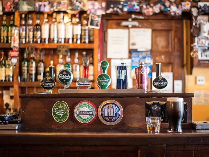 En bardisk med ølhaner, flasker på hylder, en halv liter mørk øl, et glas whisky og billeder på væggen i baggrunden.